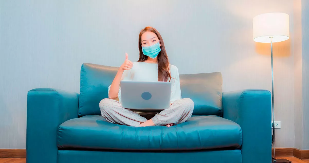 Fotografía de una chica con tapabocas sentada en un sofa con un portatil haciendo señal de aprobación con el pulgar